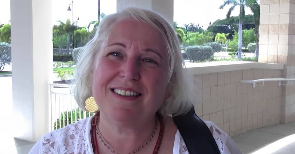 Gail smiling with platinum blonde hair wearing white lace top and gold disc earrings on a covered porch with palm trees behind