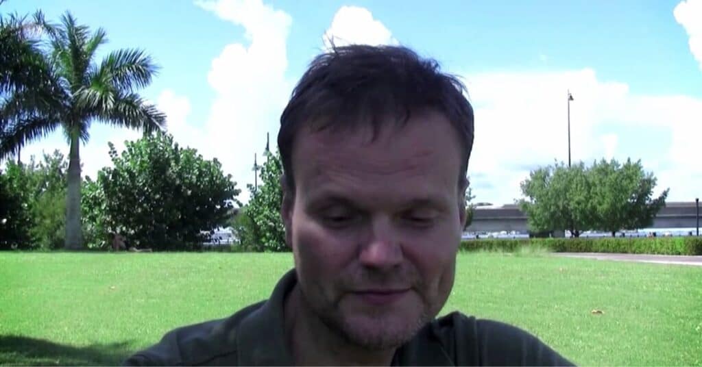 A Giedrius Kasiulynas with nearly closed eyes smiling slightly outdoors in a park with palm trees and green lawn in the background.