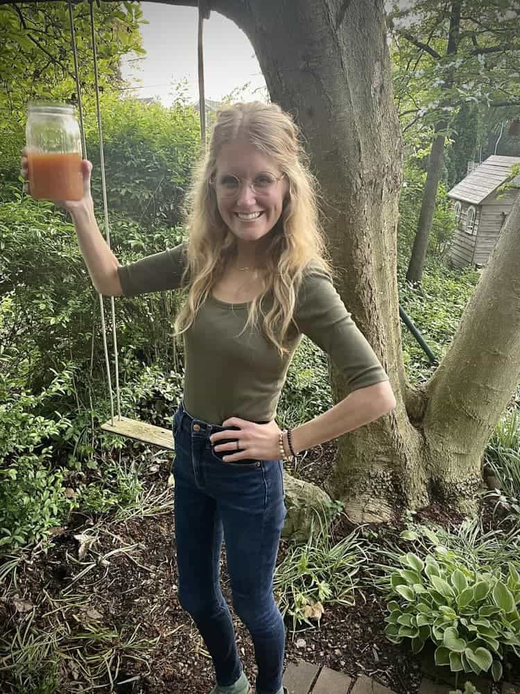 Chelsea in olive top holding up mason jar with juice in garden setting, standing near tree swing.