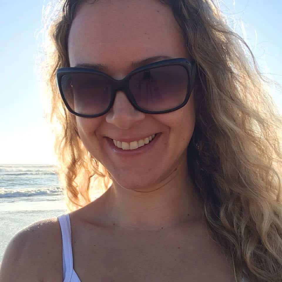 Nina smiling with wavy blonde hair wearing black sunglasses at the beach, ocean waves visible in background.