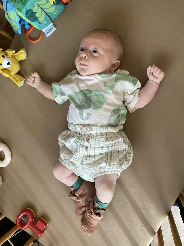 Baby lying on brown surface wearing dinosaur t-shirt and striped shorts, surrounded by toys, with green socks and brown booties.