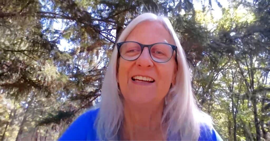 Terri smiling with long white hair and glasses wearing bright blue, photographed outdoors with sunlit trees in background