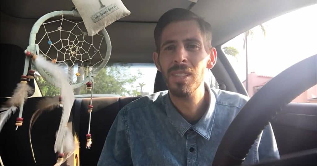 Jon in car wearing denim shirt, sitting in car with decorative dreamcatcher hanging from rearview mirror and small pillow visible