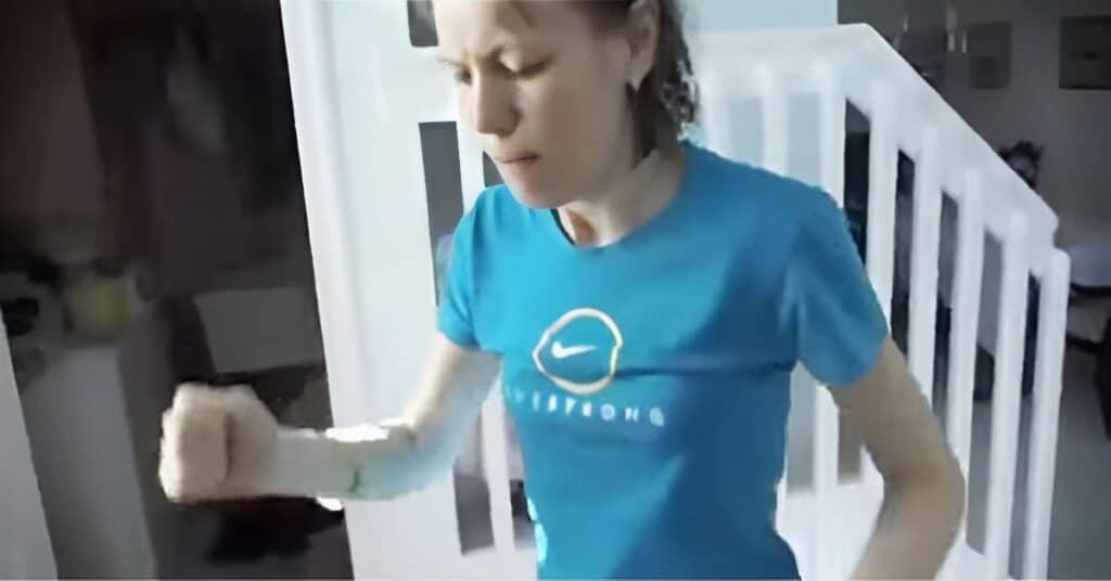 Jurgita in turquoise Nike t-shirt examining their bandaged arm near a white staircase railing in a home interior setting.