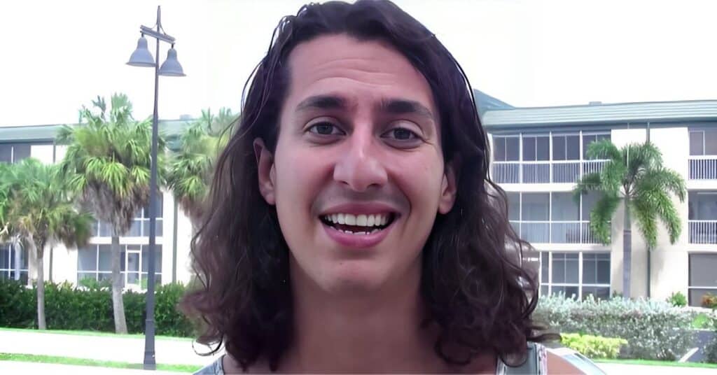 Jose smiling with long dark wavy hair outdoors, palm trees and apartment building visible in background, bright daylight.