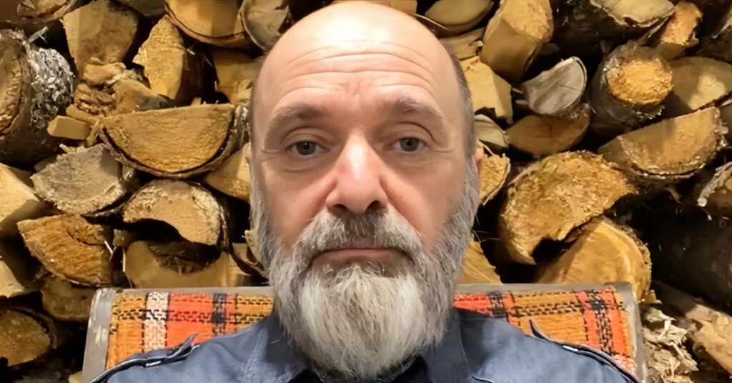 Steven sits in front of stacked firewood, wearing a blue shirt, with an orange patterned cushion visible.